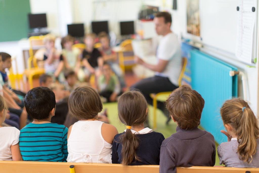 5 enfants de dos en premier plan qui écoutent leur instituteur. En arrière-plan, l'instituteur et le reste de la classe sont un peu flous.