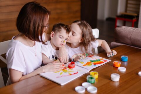 2 enfants avec leur maman ou une assistante maternelle. le petit garçon semble porteur d'un handicap. Les deux enfants font de la peinture sur un cahier posé sur la table devant eux.