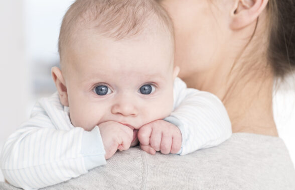 bébé aux grands yeux bleus porté par une femme brune. Le focus est fait sur le bébé uniquement. Il porte un pull blanc avec des rayures bleu clair et a les poings serrés sur l'épaule de la femme.