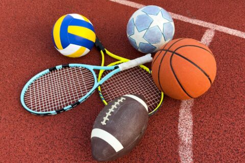 matériel de sport : ballons de football, de handball et de rugby, ainsi que raquettes de tennis posés sur la surface d'un terrain de sport