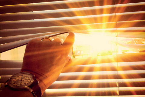 personne qui regarde l'extérieur au travers de persiennes. Grand soleil à l'extérieur. Pièce plongée dans l'obscurité