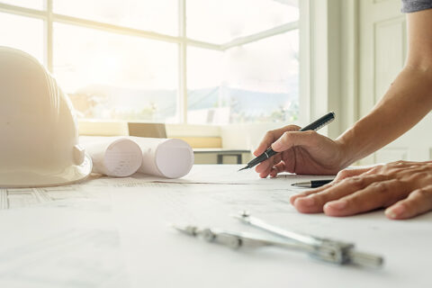 un homme qui travaille sur des plan d'urbanisme avec un casque blanc et un compas posés sur la ptable à ses côtés