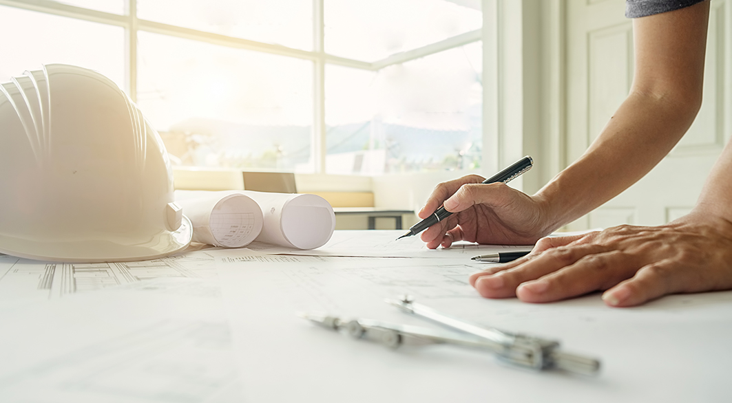 un homme qui travaille sur des plan d'urbanisme avec un casque blanc et un compas posés sur la ptable à ses côtés