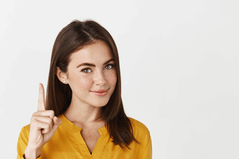 Jeune femme aux cheveux longs et bruns portant une chemise jaune. Elle lève le doit pour signifier de faire attention à quelque chose.